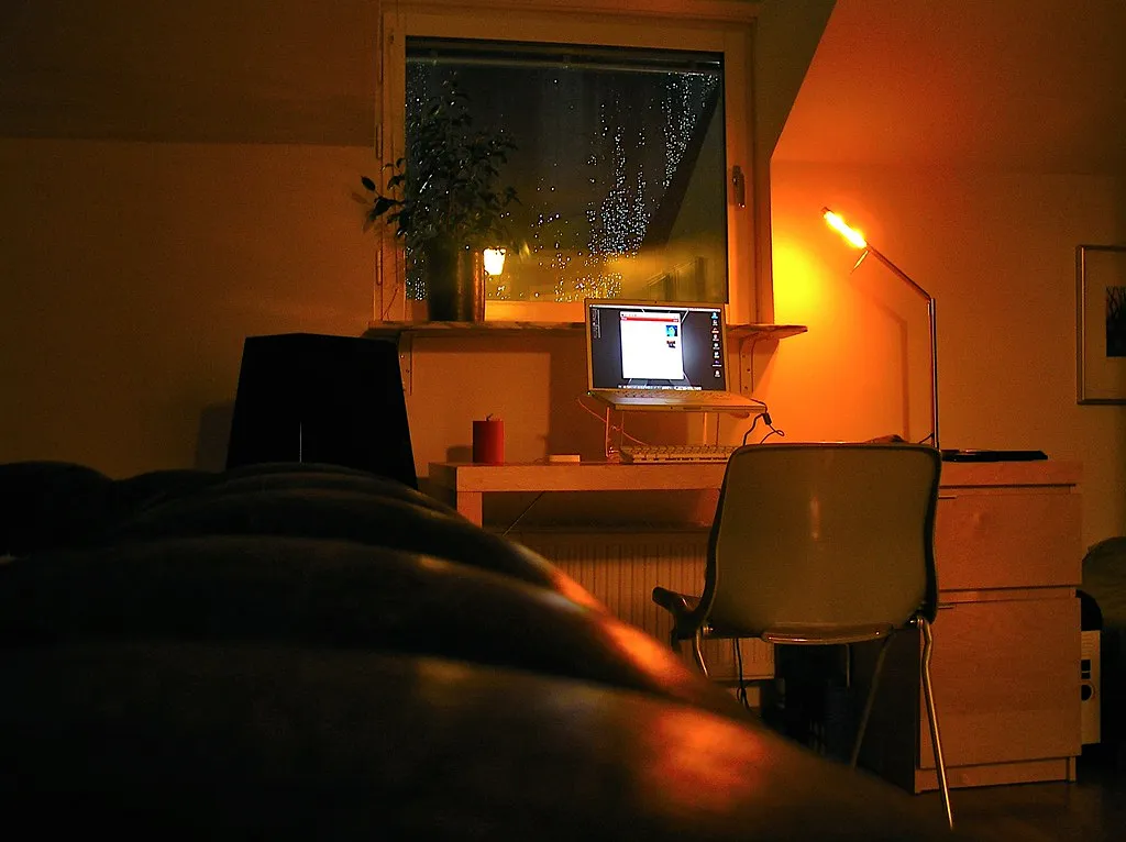 A desk with a glowing lamp and laptop beside a rain-flecked window at night