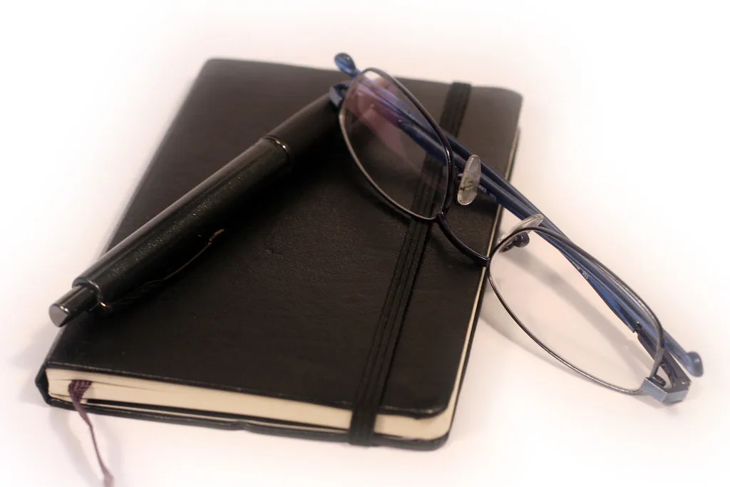 A closed notebook with a pen and reading glasses resting on top