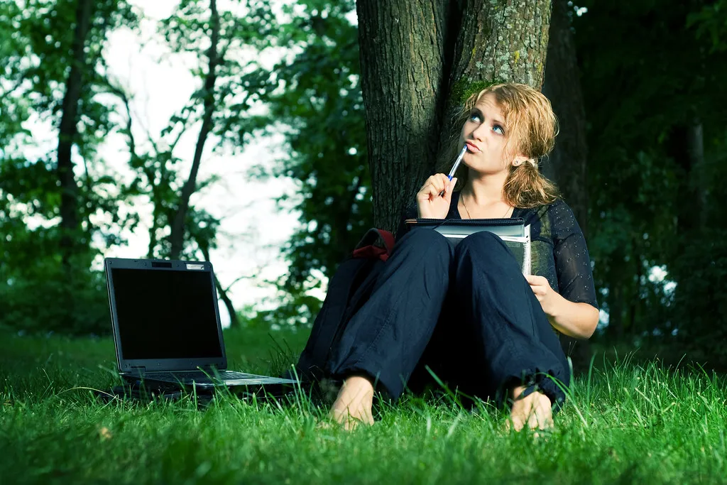 A student sitting under a tree with a notebook and a laptop in the grass